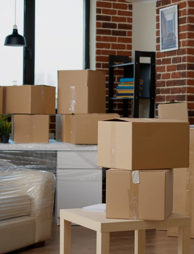 Nobody in empty living room with cardboard packaging at new home, furniture things in stack of carton boxes. No people in household property with package cargo to move in, real estate.