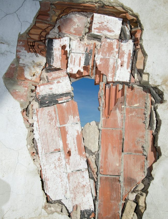 A damaged brick wall revealing a hole with a clear blue sky visible through it.